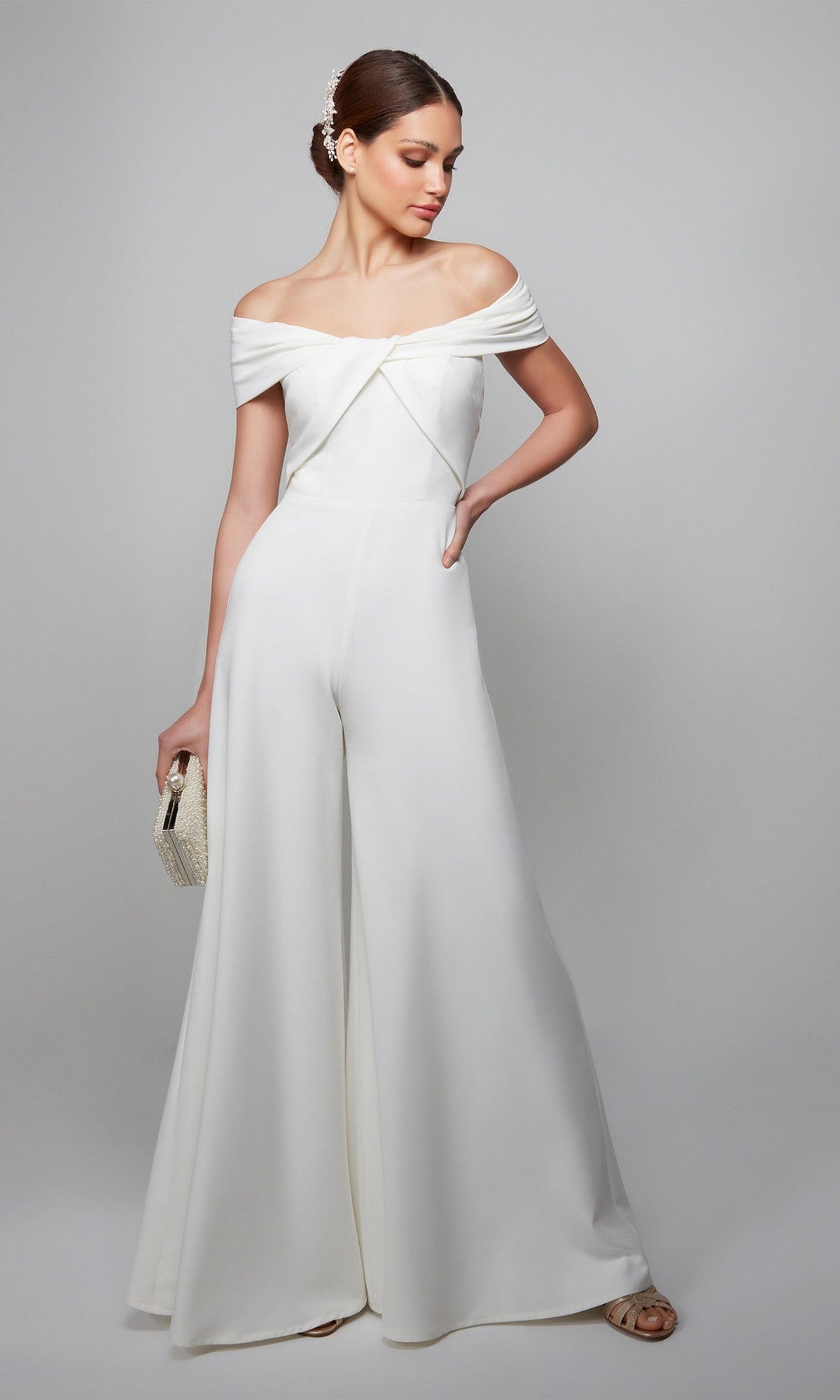 A woman in an ivory Alyce 70013 long formal jumpsuit with wide-leg pants holds a small clutch while standing against a plain light background.